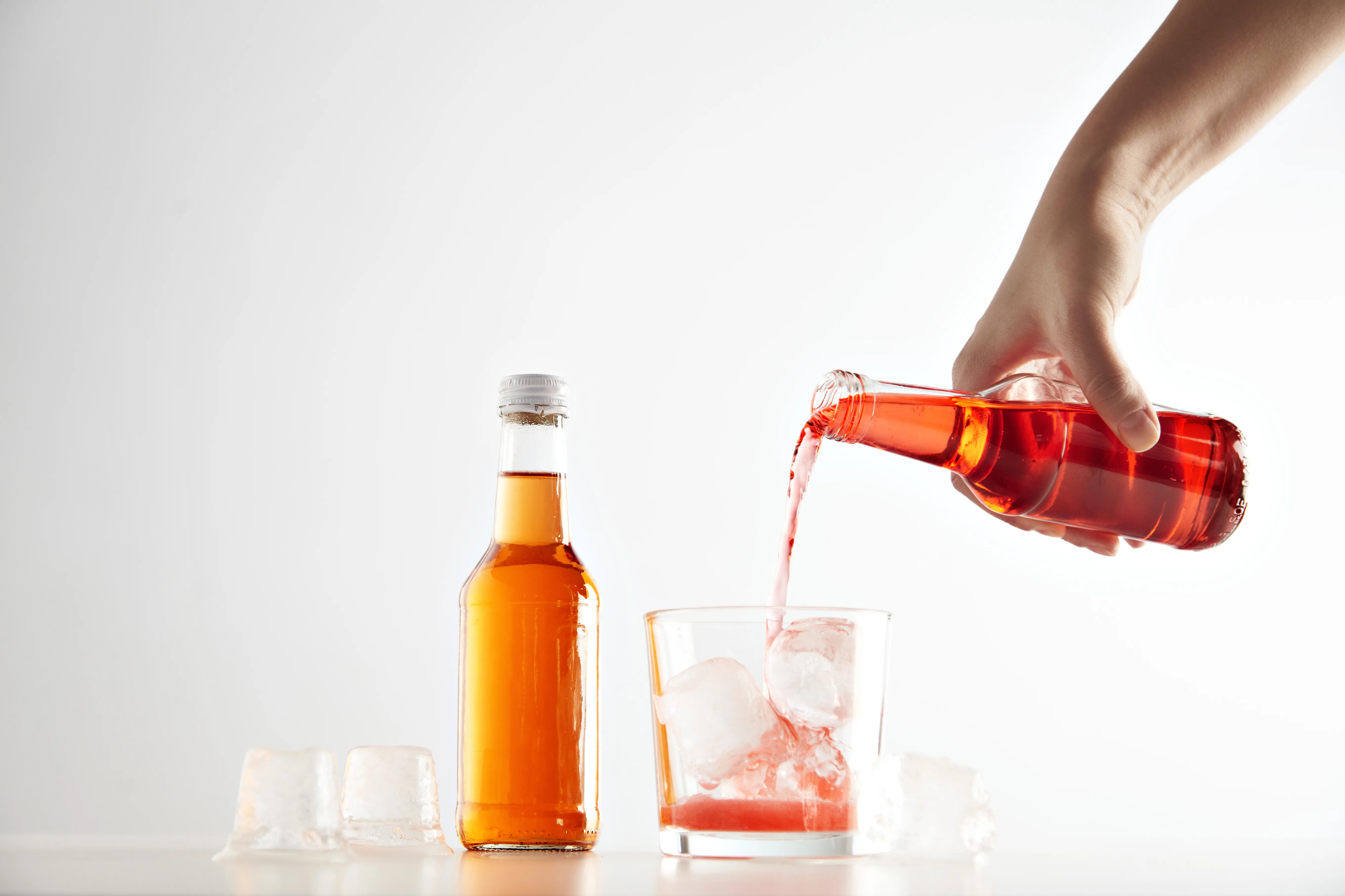 Hand pours berry cider drink into glass with ice cubes near sealed closed unlabeled bottle with orange aperol